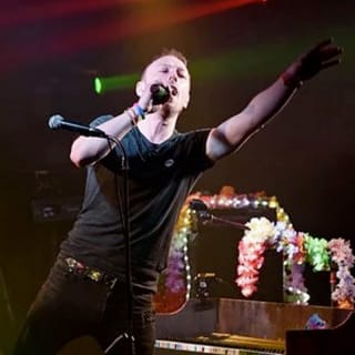 An adult male on a stage singing at a rock gig. He's got short light brown hair, a black tee and dark jeans singing into the microphone and pointing out to the crowd. image