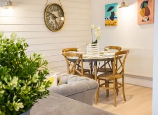 A glimpse of the dining area in one of our cottages. A four-seater round table set ready for dinner. two pictures and a clock hanging on the walls around the table. A section of a sofa is seen in the left hand corner. image