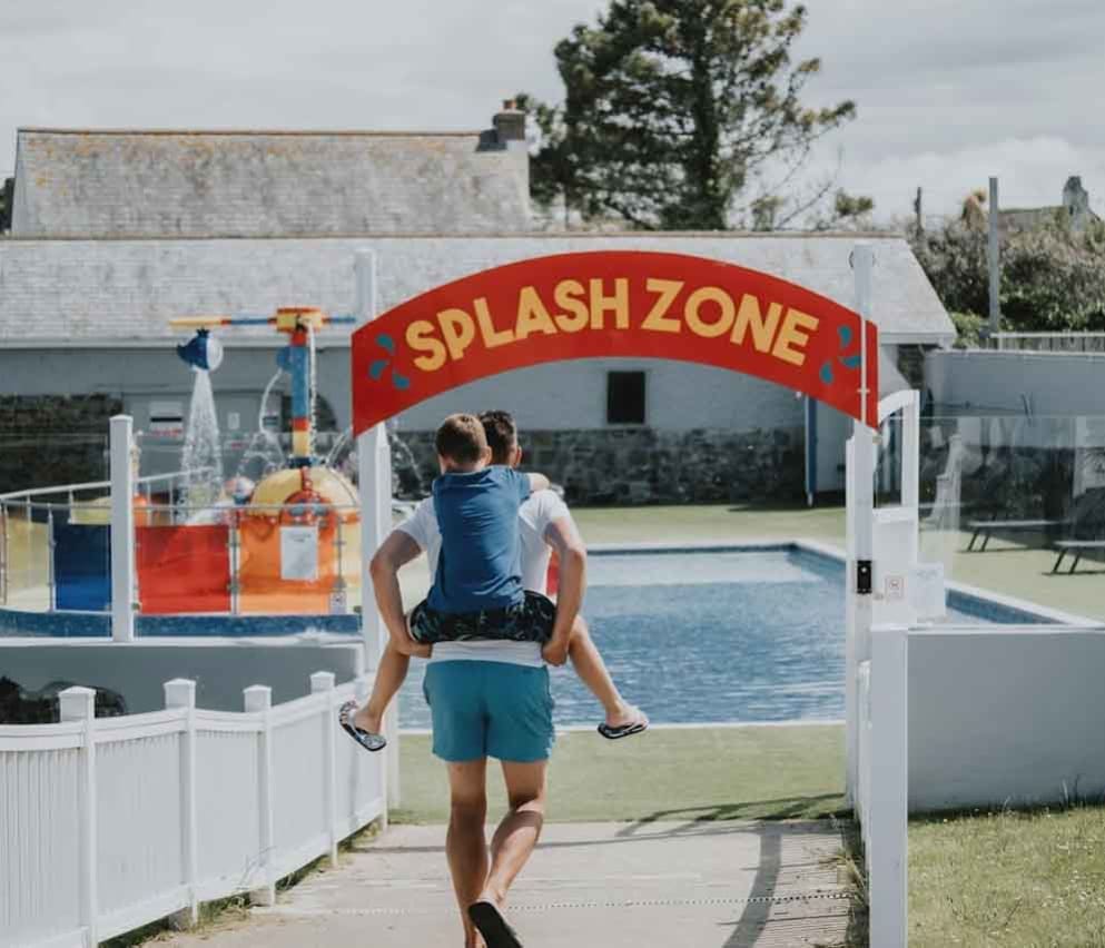 A young boy on his father's back heading towards the outside pool area. A read sign with yellow writing reads 'Splash Zone'.