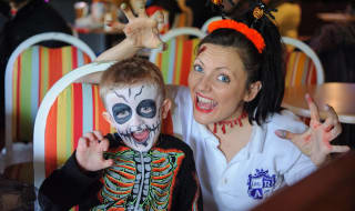 A young white boy with a skeleton face paint and outfit is holding his hand up like a claw with a white woman who is wearing face paint-style injuries to her head and neck, whilst also showing both her hands like claws. image