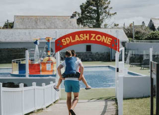 A young boy on his father's back heading towards the outside pool area. A read sign with yellow writing reads 'Splash Zone'. image
