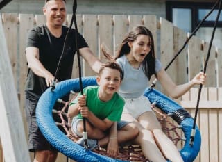 Two children playing on the swing at our adventure playground at Bude Holiday Park. There is a male pushing the swing. image