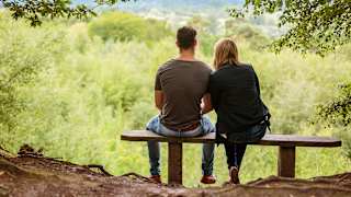 A couple sat on a bench looking out to stunning woodland views on a summers day.