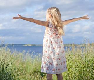 A young girl, with mid-length blonde hair, has her arms up in the air open wide, whilst looking out to sea. She is standing next to long grass, with the view of the sea in the background.
