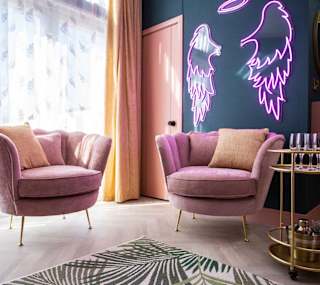 The stunning lounge area in The Esme Compact. There are two plush velvet chairs, a gold drinks trolley and some pink neon lighting in the shape of angel wings. image