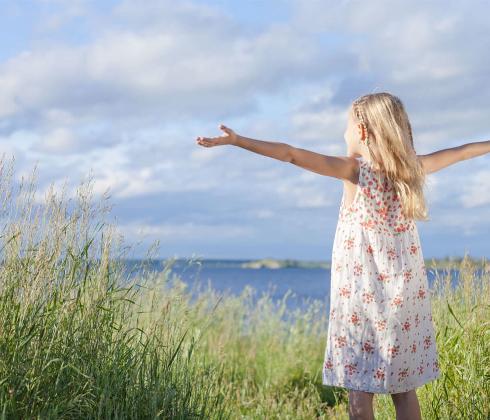A young girl, with mid-length blonde hair, has her arms up in the air open wide, whilst looking out to sea. She is standing next to long grass, with the view of the sea in the background.