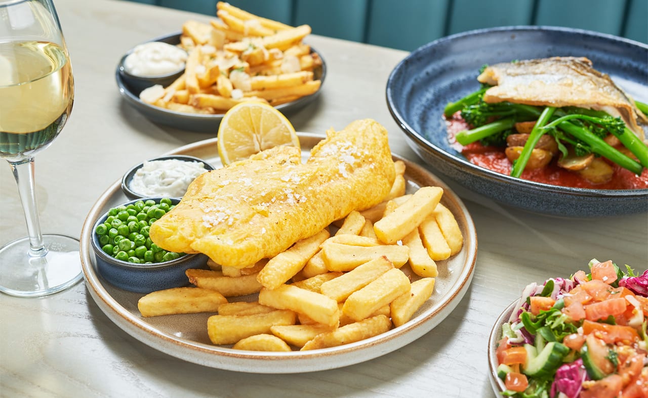 A close up of fish and chips on a wooden table with various other dishes around it. image