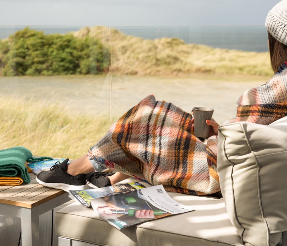 A lifestyle image of a woman relaxing on the decking at our stunning holiday park in Cornwall, St Ives Bay. There are views of the ocean in the background.