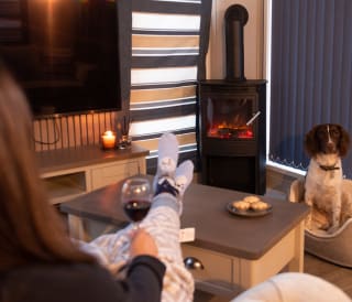 A lifestyle image of a guest cosying up by the fire in one of our stunning holiday homes. She is holding a glass of mulled wine and there are some mince pies on the table. A dog can be seen in the background in it's cosy bed.