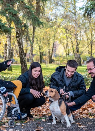 two women and two men are pictured with a medium sized dog. One woman is in a wheelchair and the others are crouched down petting the dog.