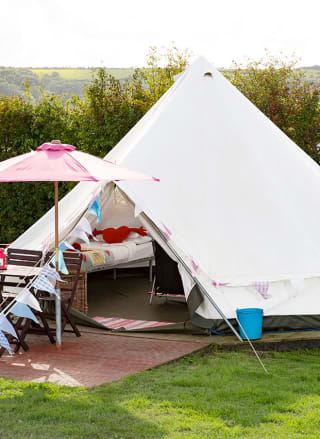 An off white canvas bell tent pitched up outside on the grass on a sunny day. The entrance to the bell tent is open and there is a pink parasol resting against the entrance.
