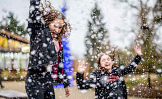 Two girls with long brown hair wearing navy blue coatsare jumping in the air excitedly and laughing as the heavy snowfall lands around them. image