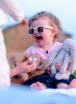 A photo of a young child with blonde curly hair tied up in a ponytail wearing white sunglasses, a pink cardegan and a navy blue dress. She is holding two rabbit teddy bears and is being offered a white dummy. The child's expression is of glee.