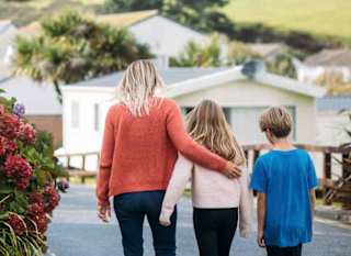 A woman with blonde hair wearing an orange jumper and jeans has her arm around a girl with light brown hair, who is wearing a pale pink top and black trousers. They are walking together down a path at our holiday park, Newquay Bay, Cornwall. image