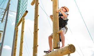 A pre-teen child is climbing on a wodden structure with a bright orange helmet, torso harness and a pully rope system. The child is looking up to judge his next move as he accends higher on the structure.