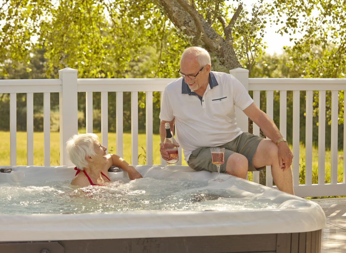 A couple lounging in the hot tub at The Lakes Rookley. The female is sat in the hot tub, whilst the male is sat alongside the hot tub. They are enjoying a bottle of wine together. image