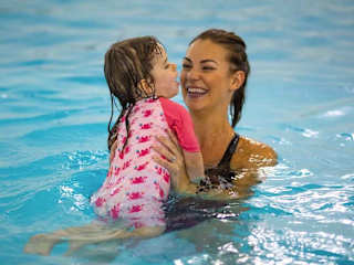 A mother and child are playing in an indoor pool. The mother is lifting the child out of the pool and the child is laughing.