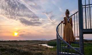 A photo of a girl with blonde hair in a bun wearing a orange polkadot dress, whom is standing in the middle of a spiral staircase looking out at the sunset.