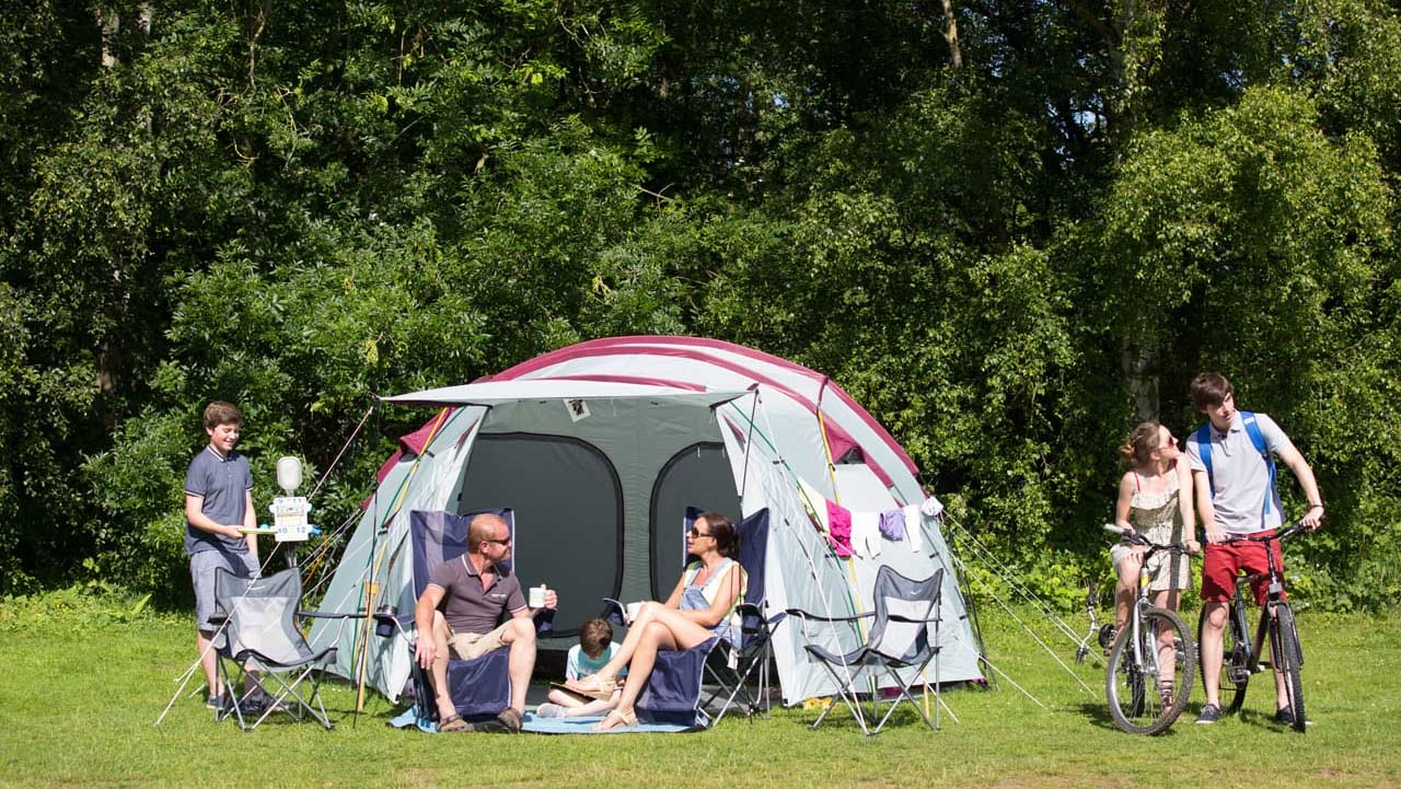 A pitched up family tent with a man, woman and baby sitting in the porch area of the tent. A teen boy and girl are cycling by on a hot sunny day.