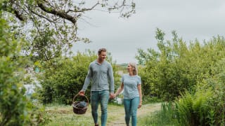 A young white couple walk hand in hand down a country path whilst the male holds a picnic basket.