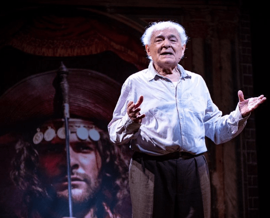 ator de 92 anos no palco, em pé de camisa branca e calça marrom