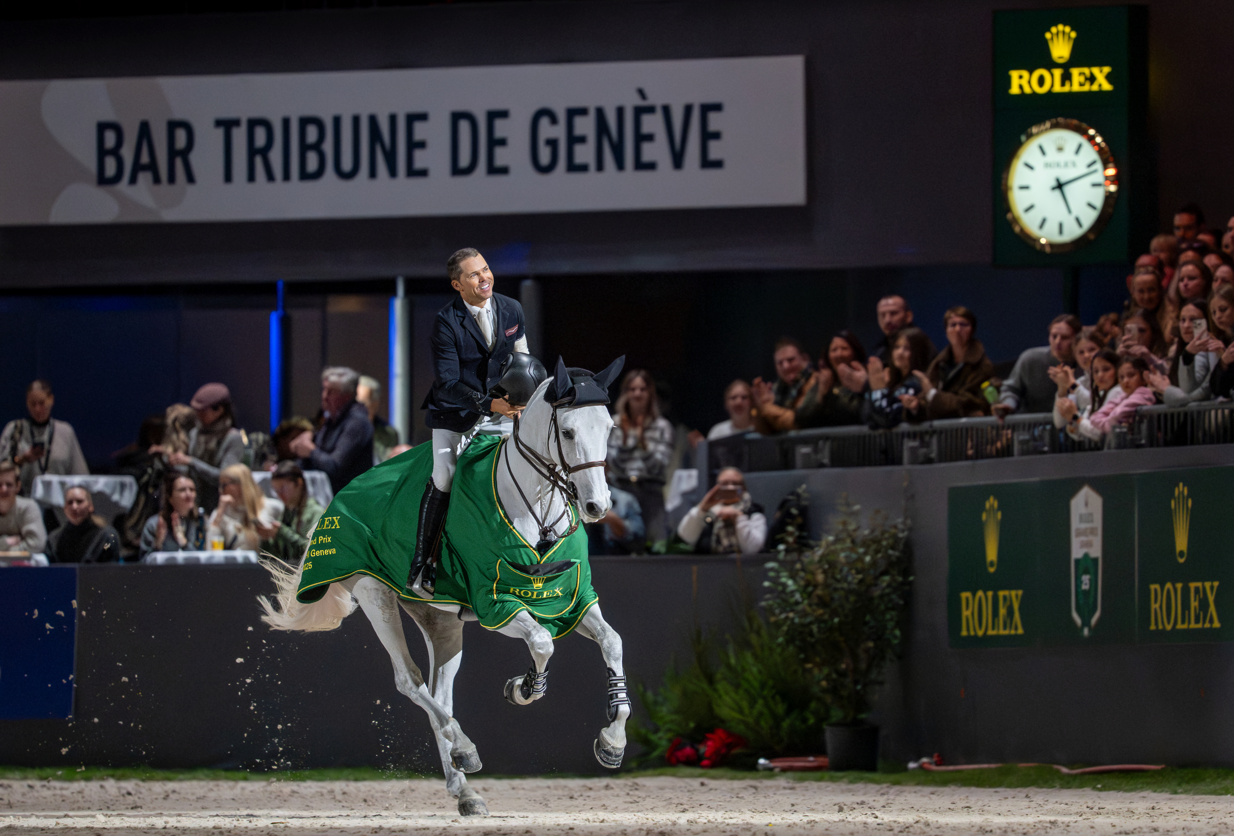 Show jumping rider galloping a grey horse in a green saddle pad through an indoor Geneva arena