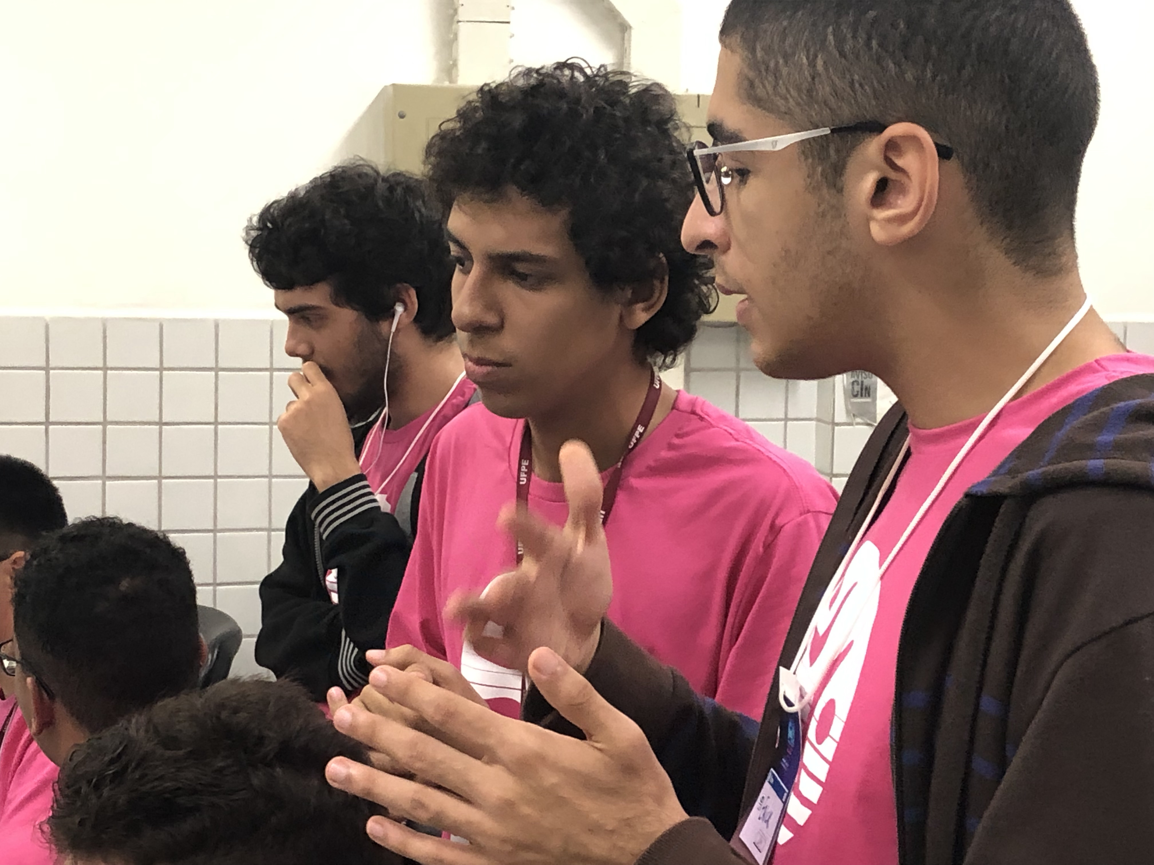 Em primeiro plano, duas pessoas conversando. Ao fundo, pessoas concentradas. Todas as pessoas estão vestindo a camisa do HackaPET.