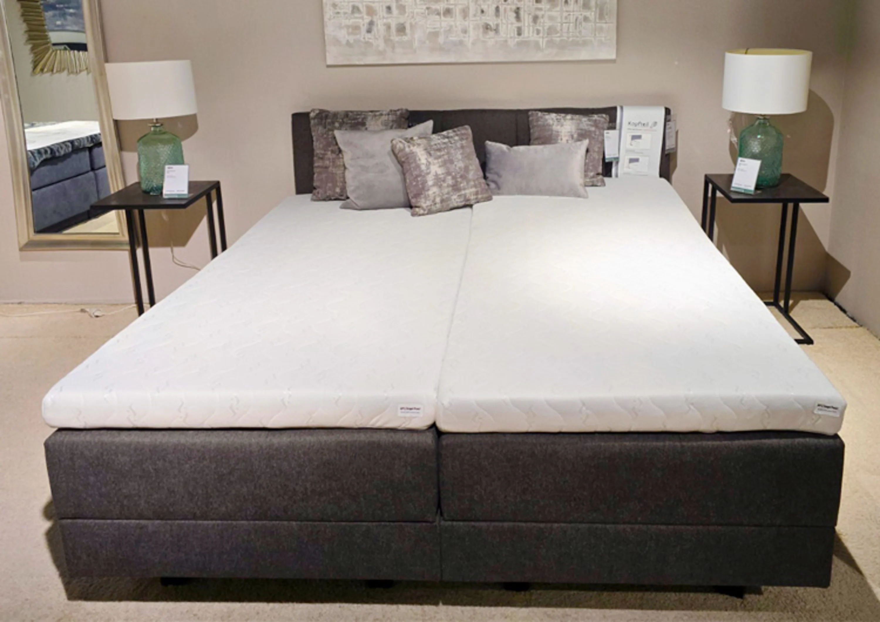 Das Bild zeigt ein modernes Boxspringbett in einem Ausstellungsraum. Das Bett besteht aus zwei Matratzen mit weißen Auflagen und einem schwarzen Untergestell.