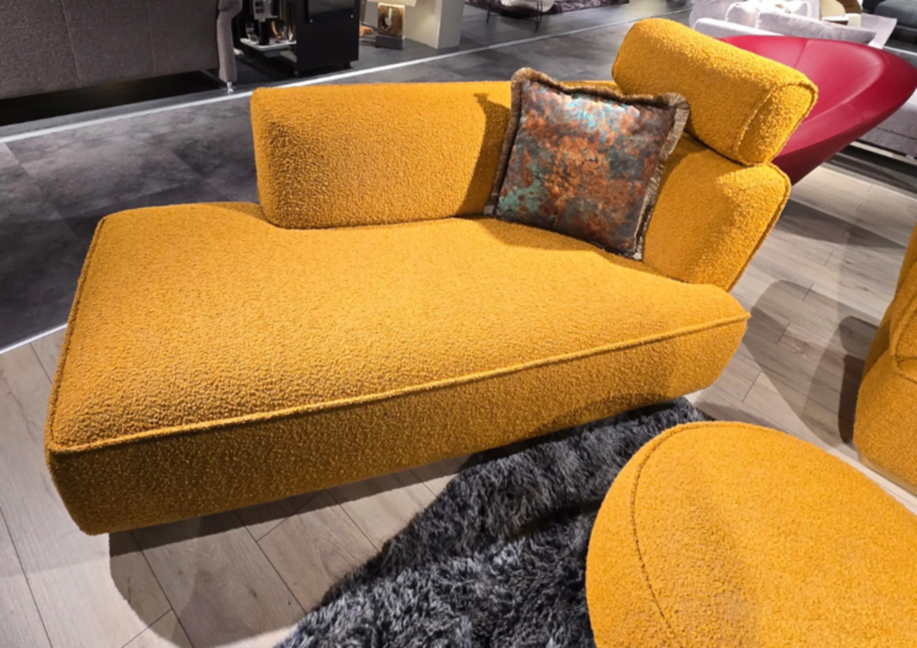 Eine moderne, senfgelbe Chaiselongue aus einem strukturierten, bouclé‑artigen Stoff zu sehen. Sie hat eine breite Liegefläche, eine seitliche Rückenlehne und eine gepolsterte Kopfstütze.