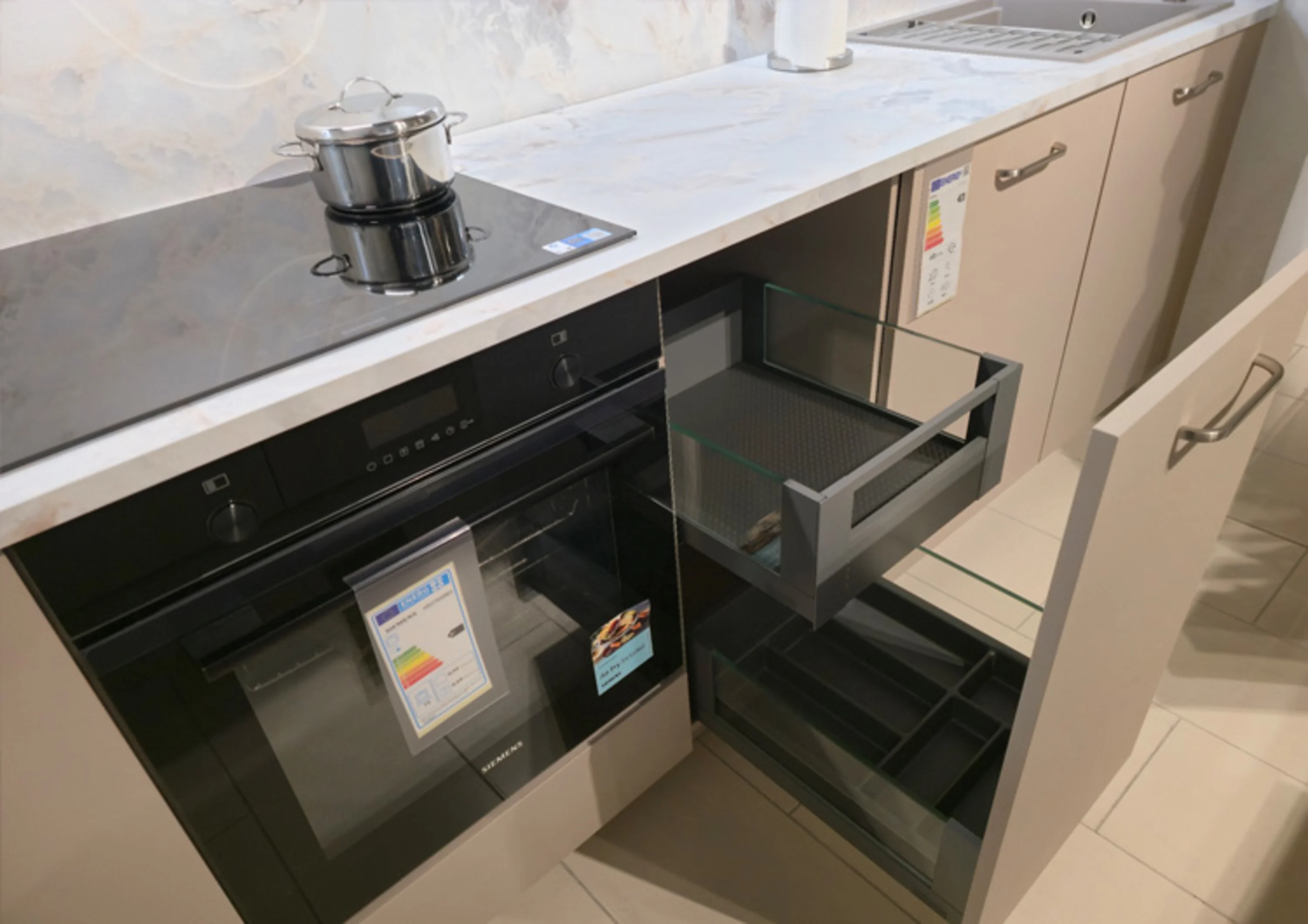 Das Bild zeigt eine Küchenzeile mit heller Arbeitsplatte in Marmor-Optik. Links sind ein schwarzer Einbau-Backofen und ein Induktionskochfeld mit einem Topf zu sehen. Rechts daneben befinden sich geöffnete Schubladen mit Glas-Seitenteilen.