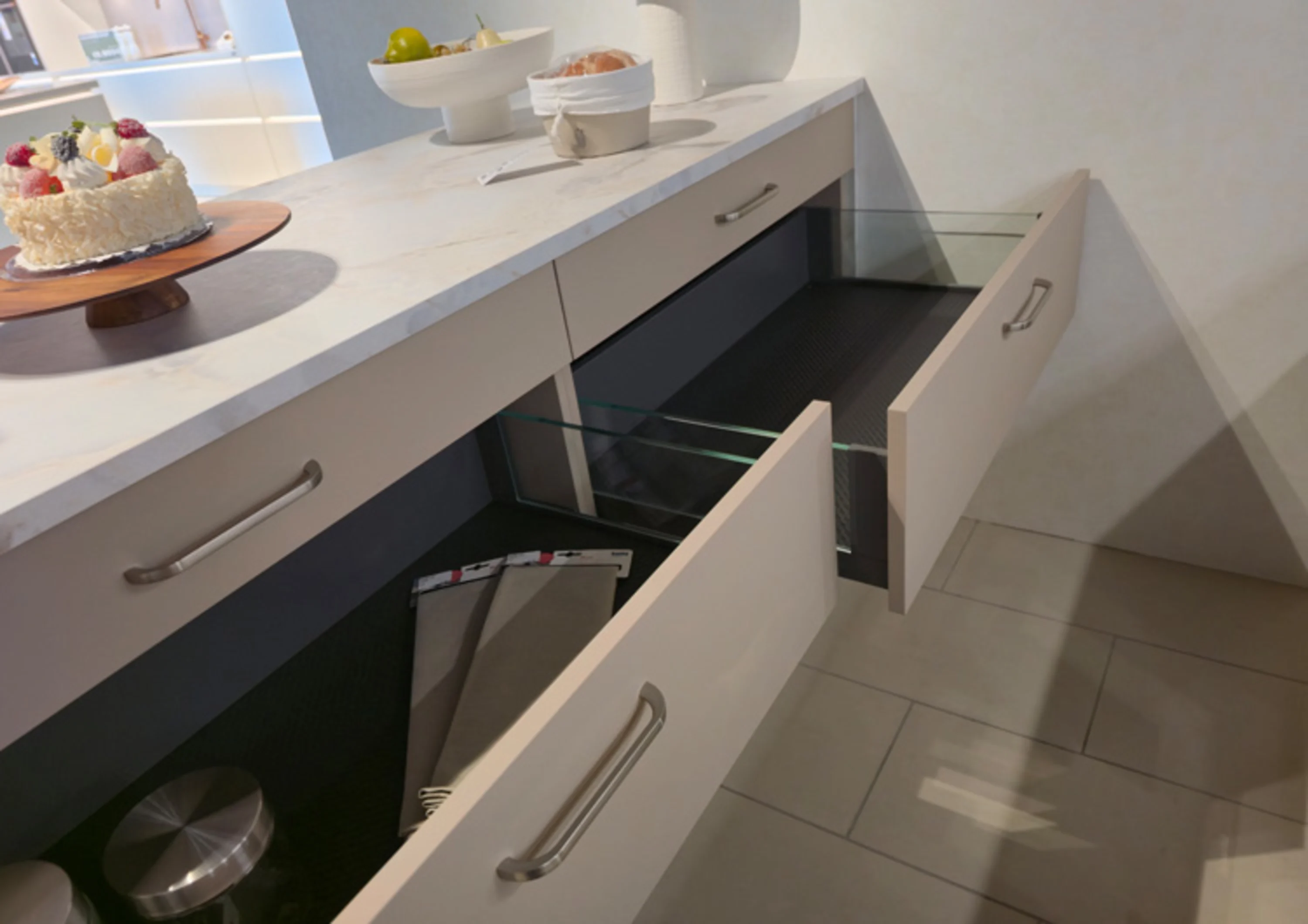 Das Bild zeigt eine moderne Küchenzeile mit einer hellen Arbeitsplatte in Marmor-Optik. Zwei breite Schubladen sind geöffnet und haben Glas-Seitenteile sowie Metallgriffe.