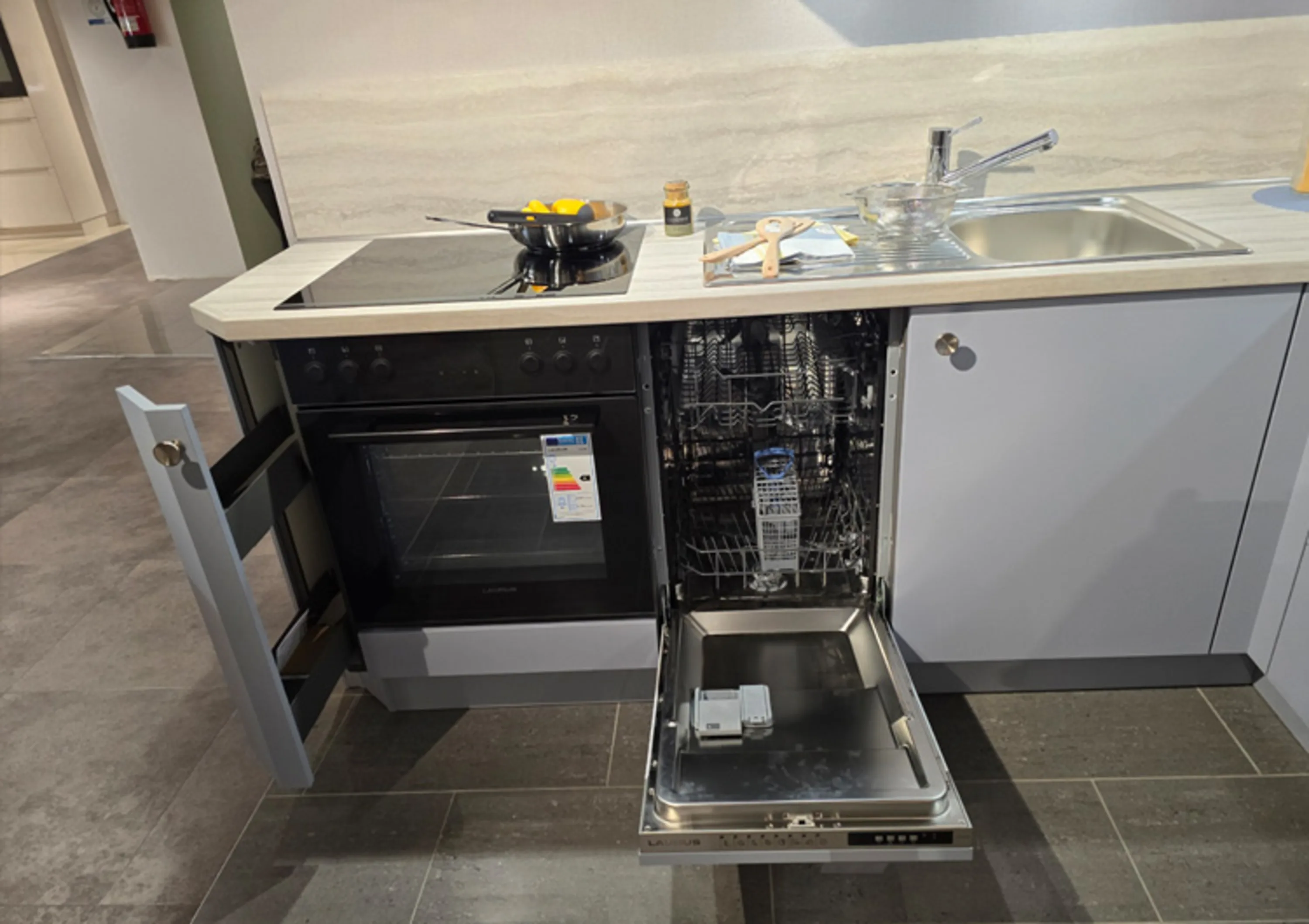 Unter der Arbeitsplatte sind mehrere Einbaugeräte zu sehen: links ein geöffneter Schrank, daneben ein schwarzer Backofen, und rechts davon eine geöffnete Geschirrspülmaschine mit sichtbaren Körben und Spülmittelbehälter.