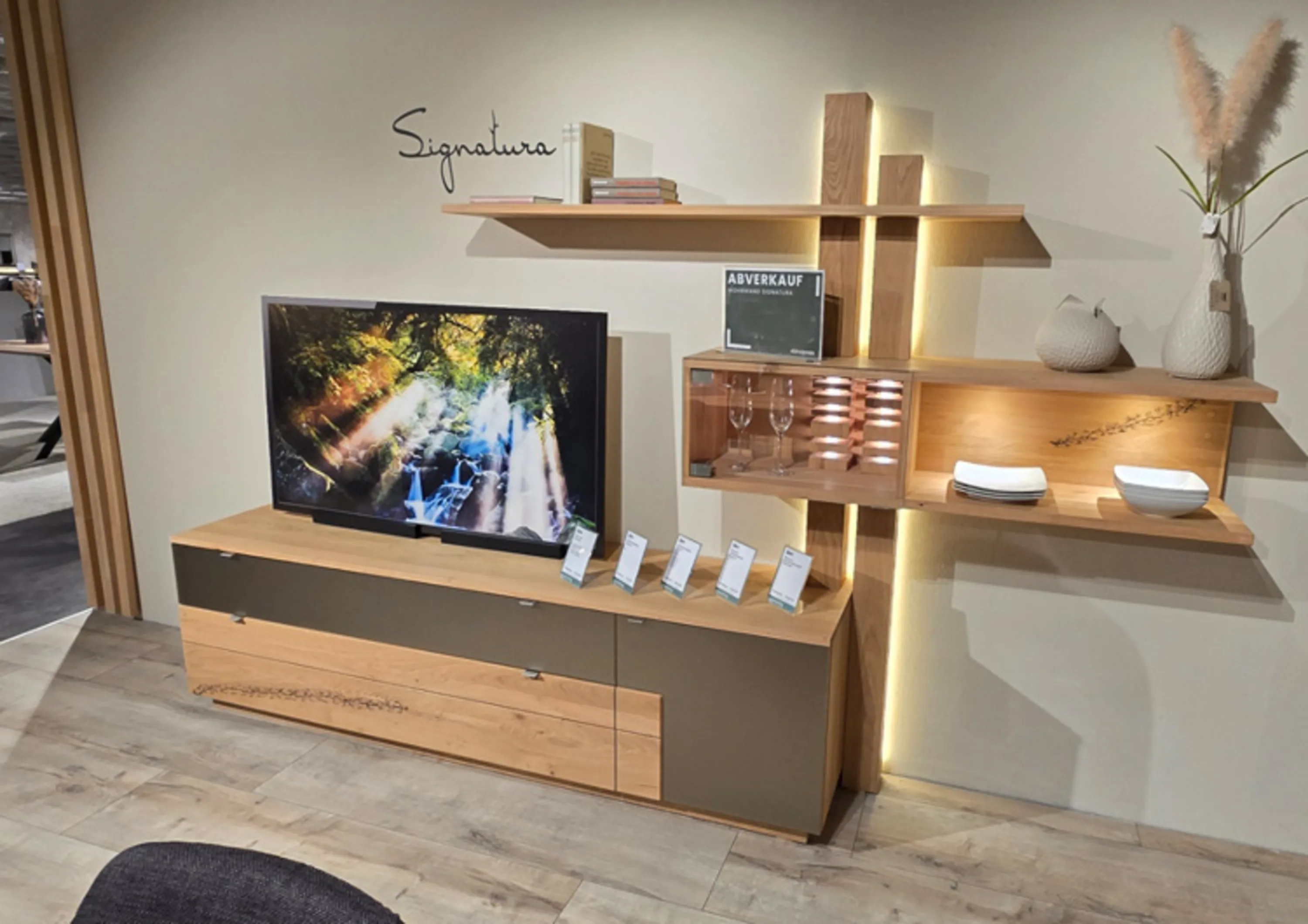 Moderne Wohnwand bestehend aus einem niedrigen TV‑Board und wandhängenden Regalen in natürlicher Holzoptik mit beleuchteten Elementen, die dekorative Gegenstände und einen Fernseher stilvoll präsentieren.