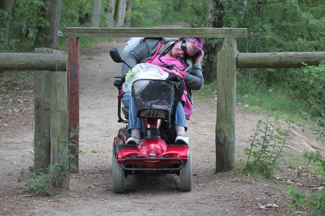 Woman in red wheelchair with bags in her lap is needing to duck under a low park entry barrier