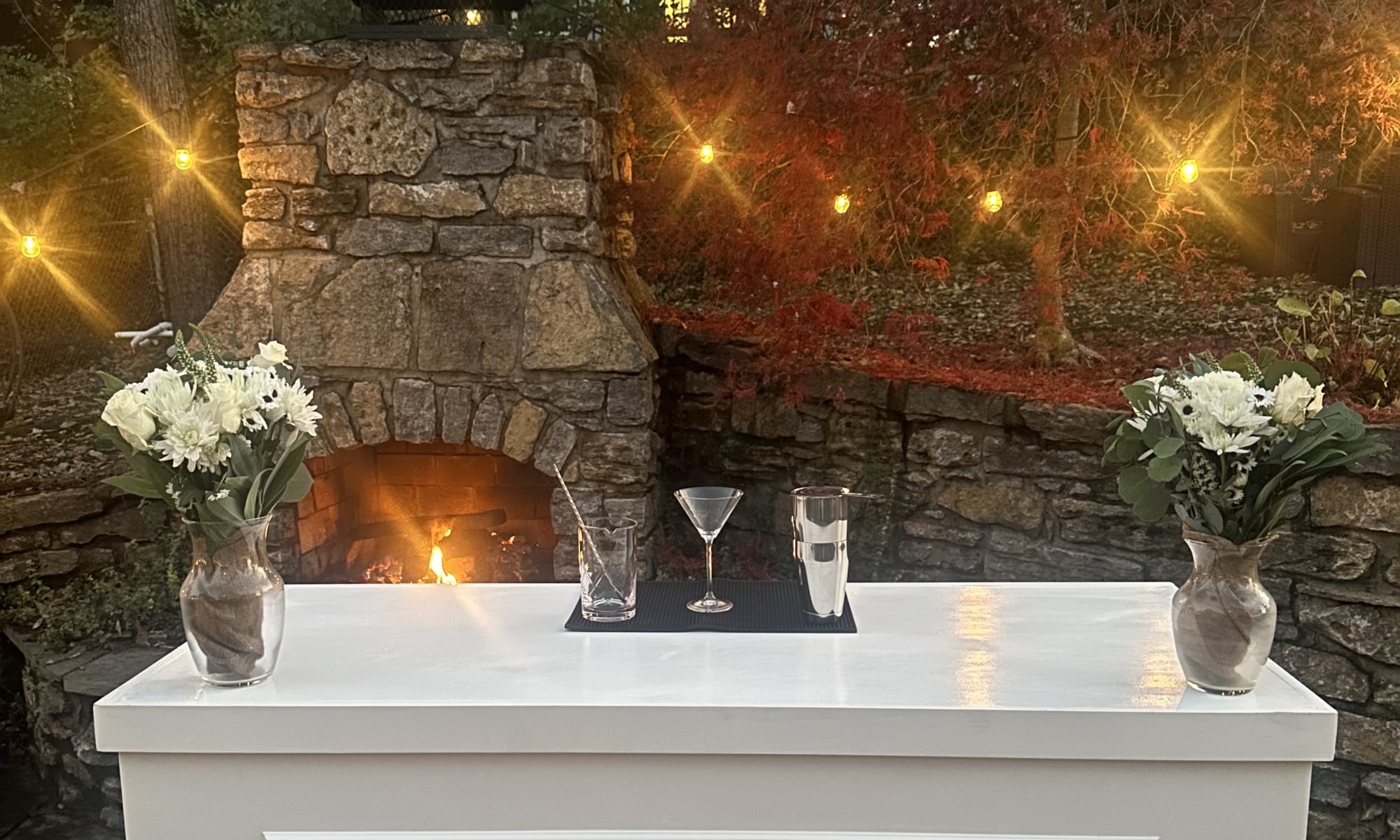 A dressed 513Sips mobile bar at night in front of a lit stone fireplace, with white florals and coupe glasses on a tray
