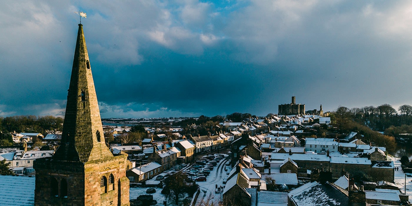 Aerial view of snow-covered Warkworth in winter — St Lawrence's church spire in the foreground, the medieval town's snowy rooftops, and Warkworth Castle on its headland in the distance under a stormy slate-blue sky