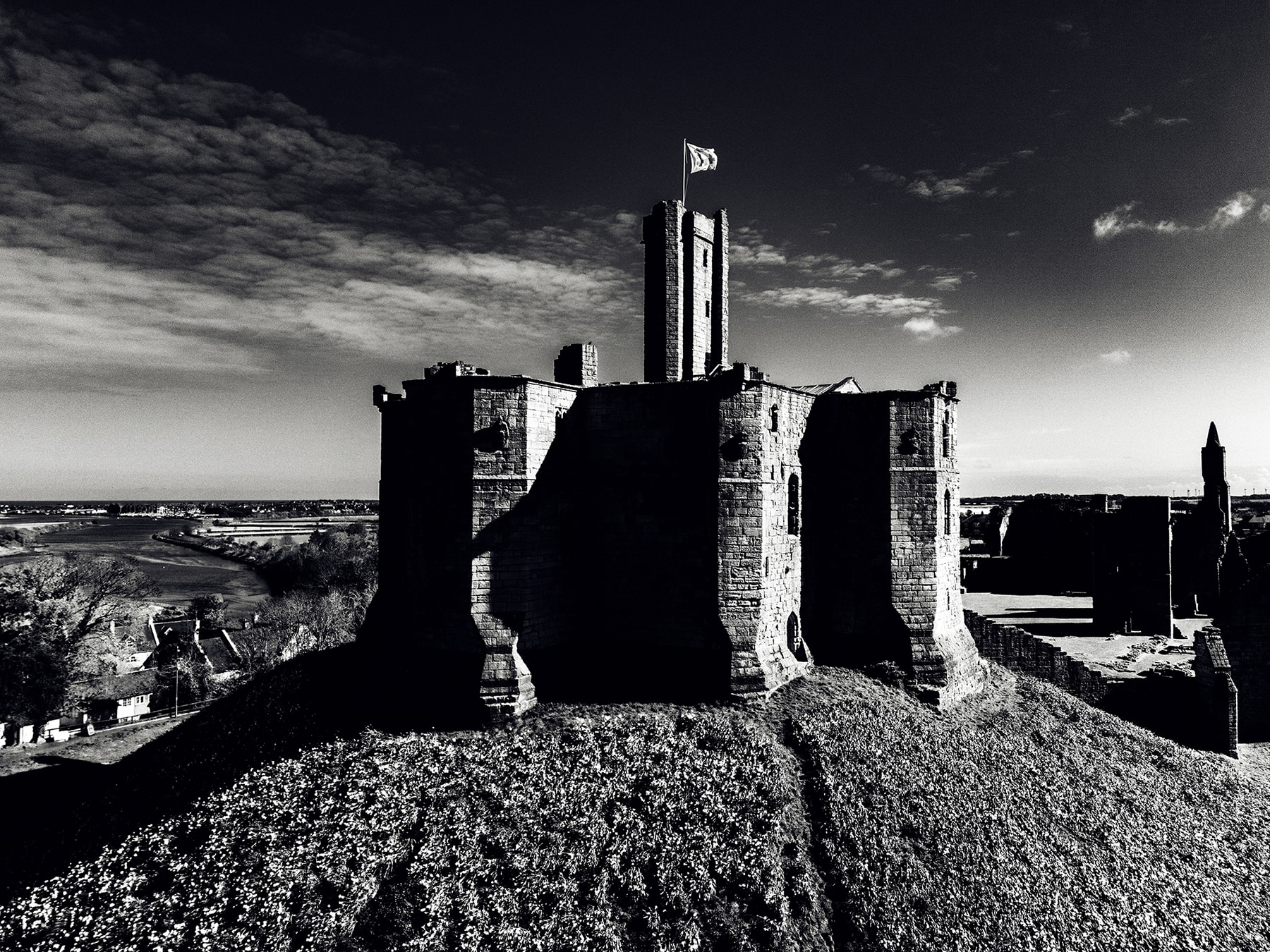 Black-and-white photograph of Warkworth Castle&rsquo;s great tower with a subtle navy tint in the shadows &mdash; the cross-shaped keep stands silhouetted, the deep blacks pick up the brand-navy cast