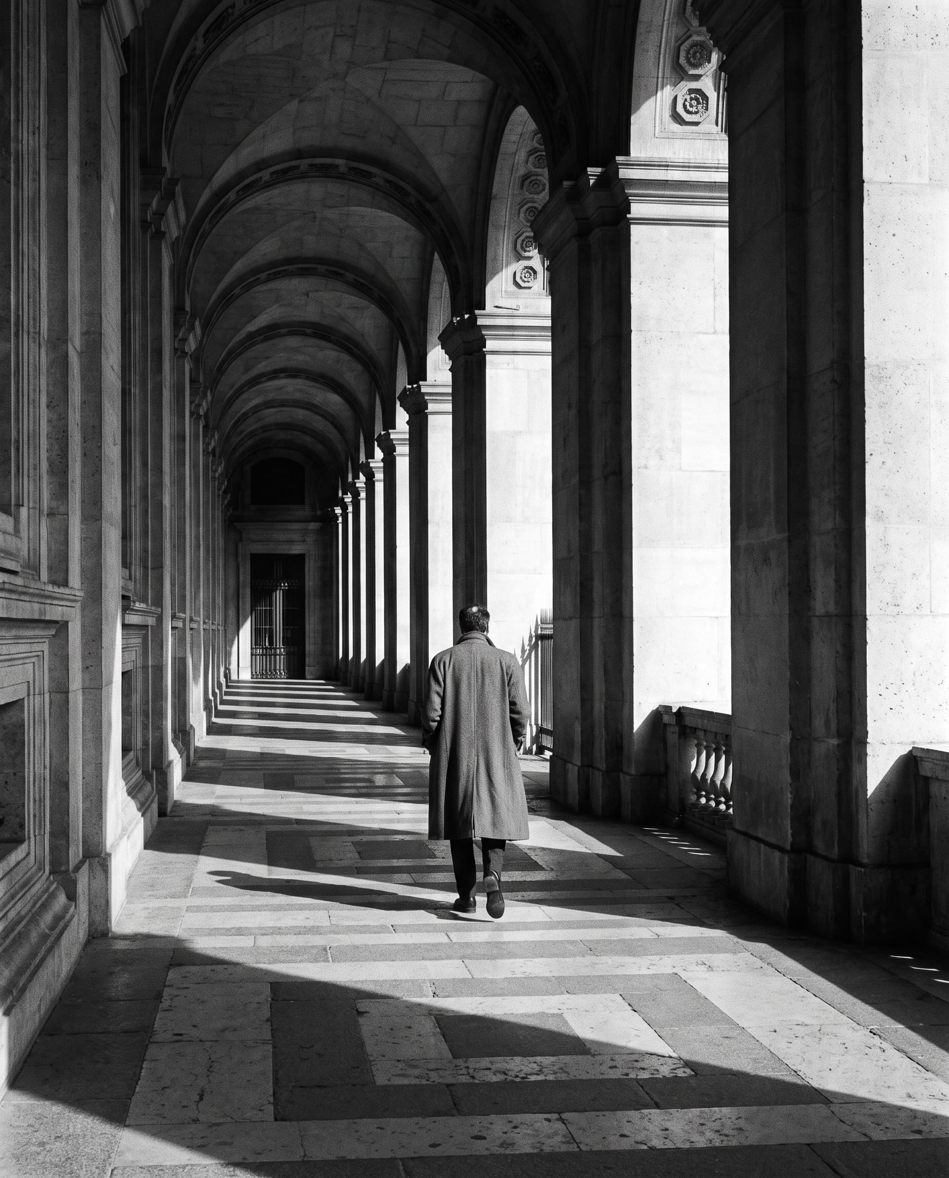 Black-and-white photo of a person walking through a long stone arcade