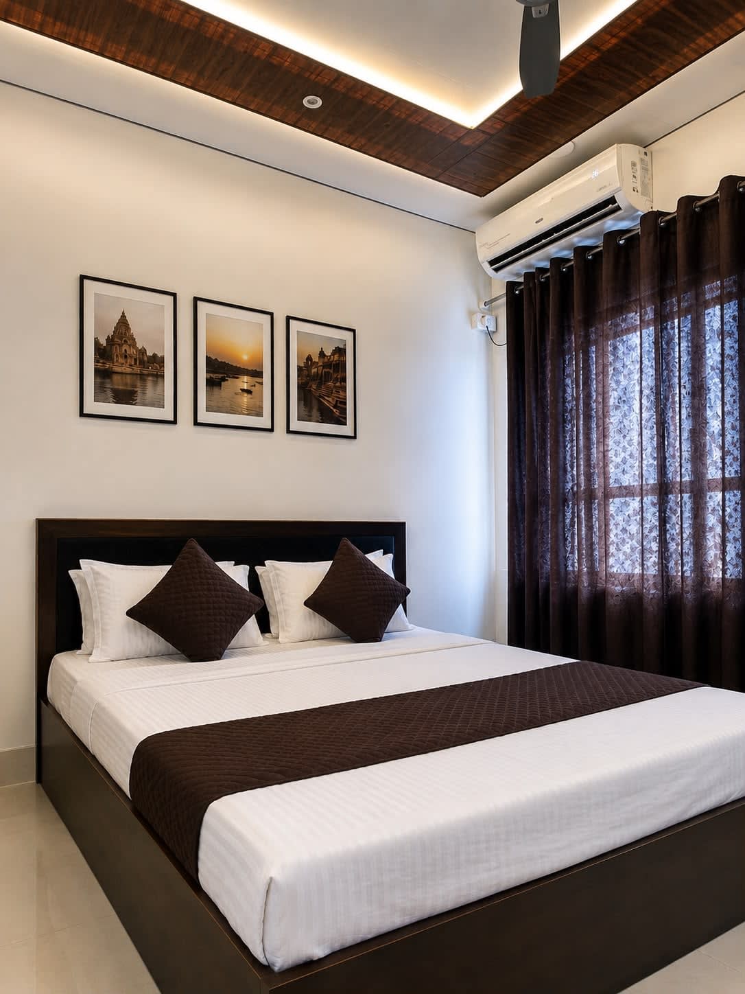 Master Deluxe Bedroom — Warm wooden false ceiling, recessed LED lighting, AC, plush double bed, and Varanasi ghat artwork
