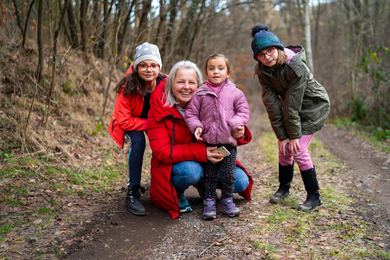 A smiling woman and three girls pause on a forest path during a chilly outdoor adventure.