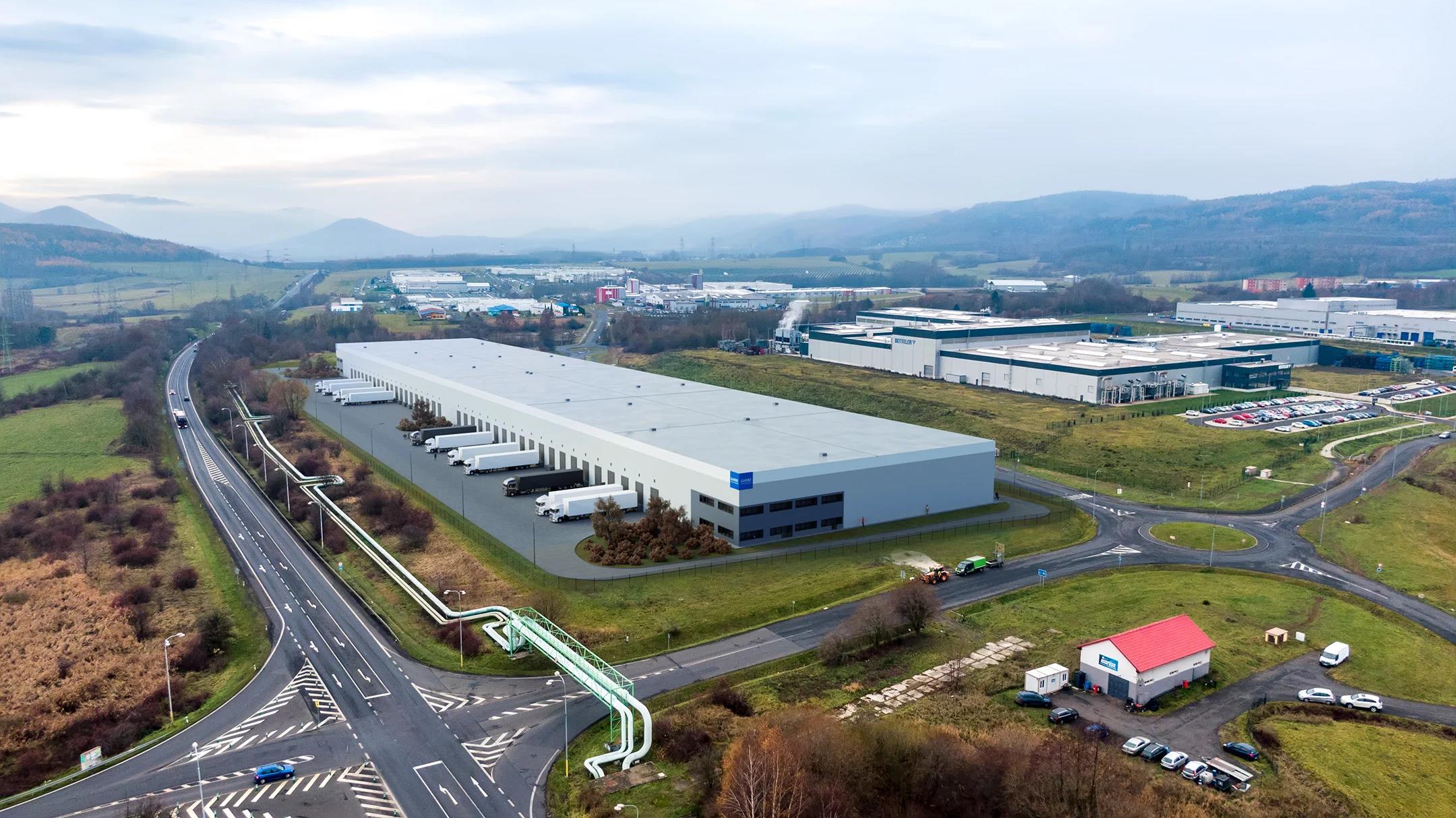 Aerial view of a large modern warehouse with trucks, roads, and mountains under cloudy skies.