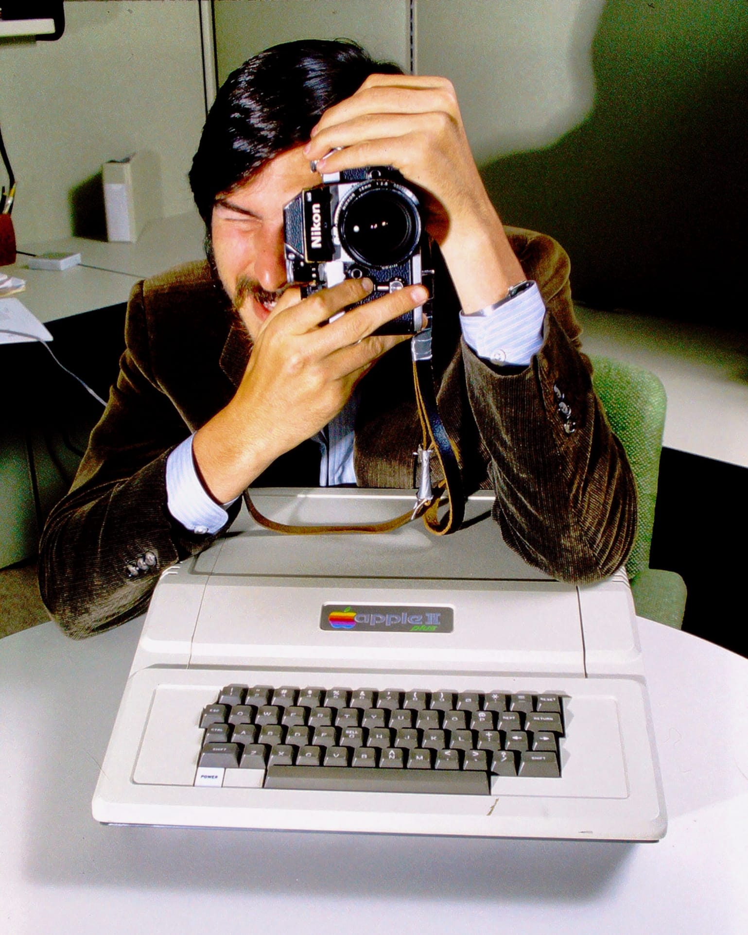 Steve in 1981, bearded and wearing a corduroy jacket, peers through the lens of a Nikon camera as his elbow rests on an Apple II. Photograph by Andrée Abecassis.