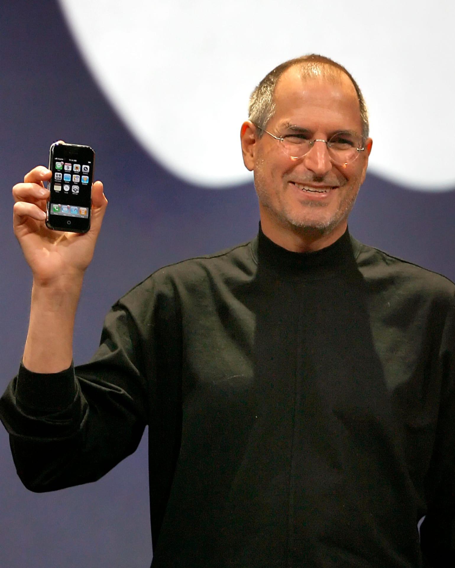 Steve in 2007, with short hair and gray in his beard, smiles and holds up an iPhone as he announces the device on stage.