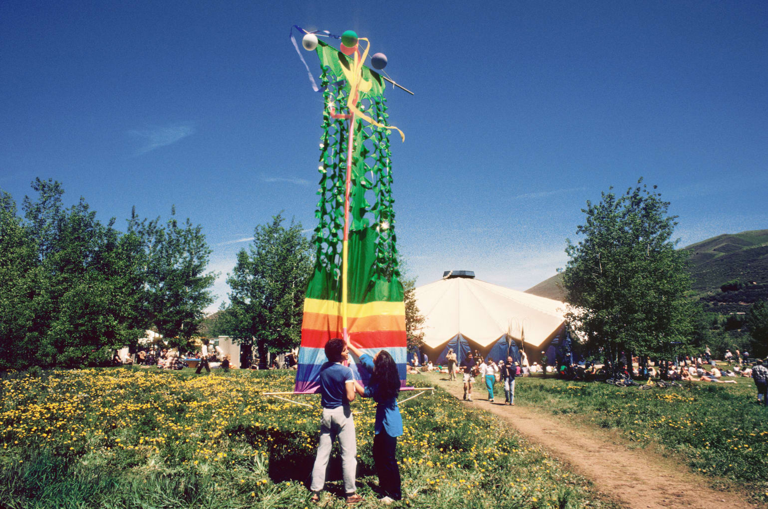 A vertical, kite-like sculpture with rainbow colors stands in a meadow of flowers, highlighting the way to the tented amphitheater.