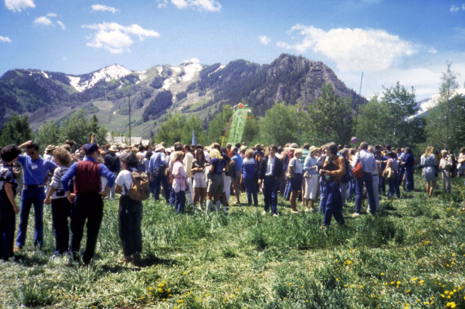 A crowd assembles on a lush, grassy field with Colorado’s Rocky Mountains (the Rockies) in the background.