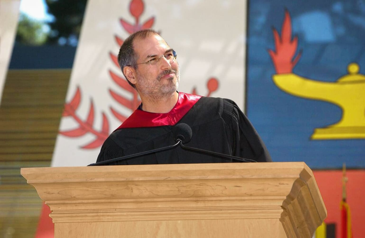 Steve Jobs stands behind a lectern and smiles at the crowd off-camera. He wears a black graduation gown with red regalia.