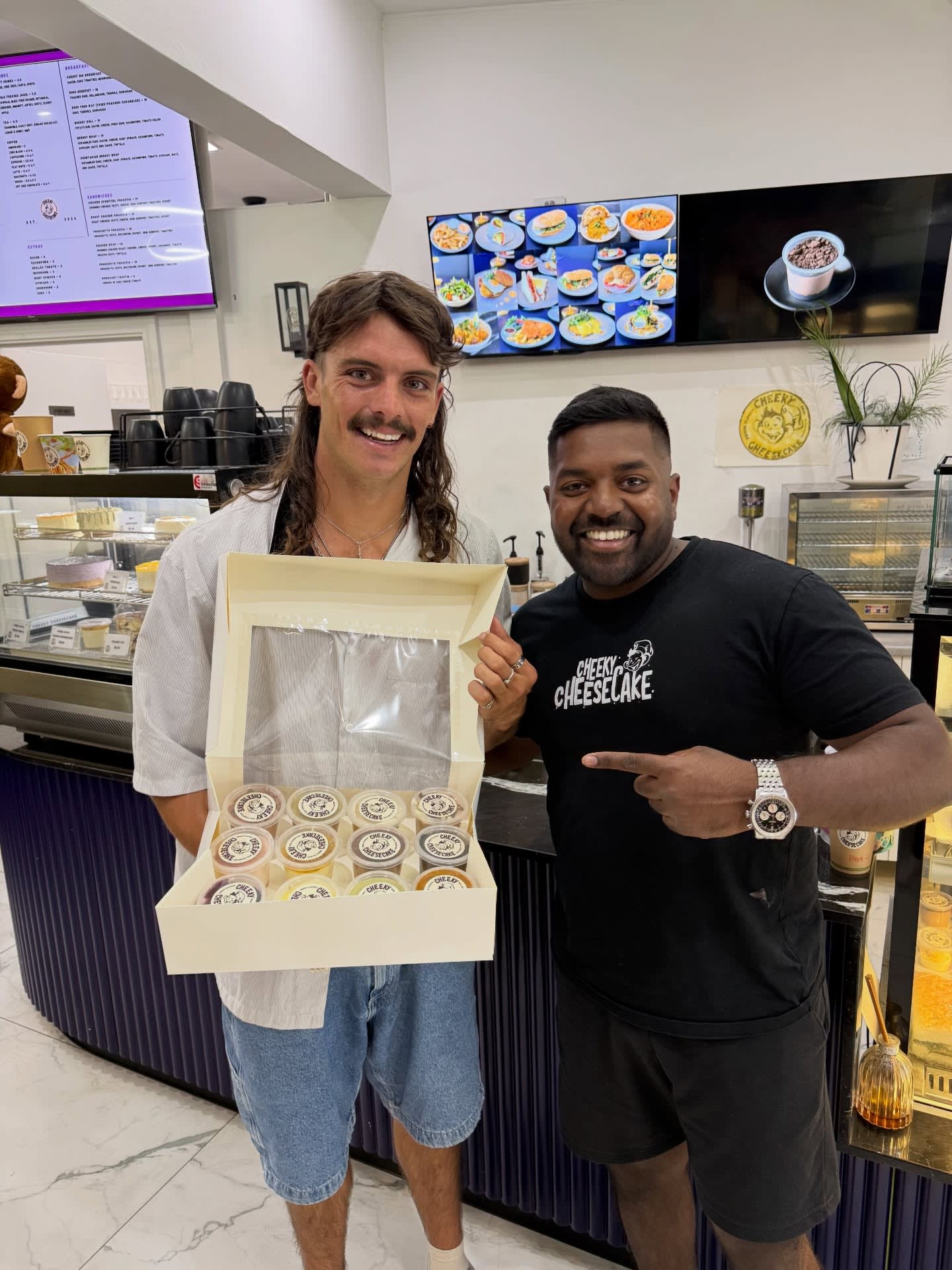 Owner handing a customer a 12-pack of cheesecake cups in front of the display case.
