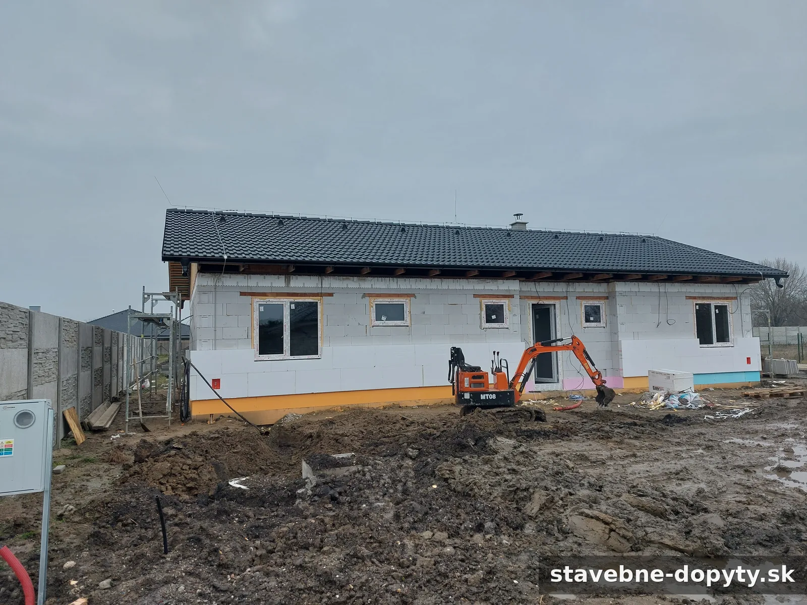 Prízemný rodinný dom vo fáze výstavby s hotovou strechou, oknami a začatým zateplením fasády, s bagrom.