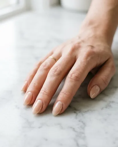 Close-up of a woman's hand with short-to-medium almond nails wearing a sheer nude gloss finish, resting on a white marble surface in soft natural light Close-up of a woman's hand with short-to-medium almond nails wearing a sheer nude gloss finish, resting on a white marble surface in soft natural light