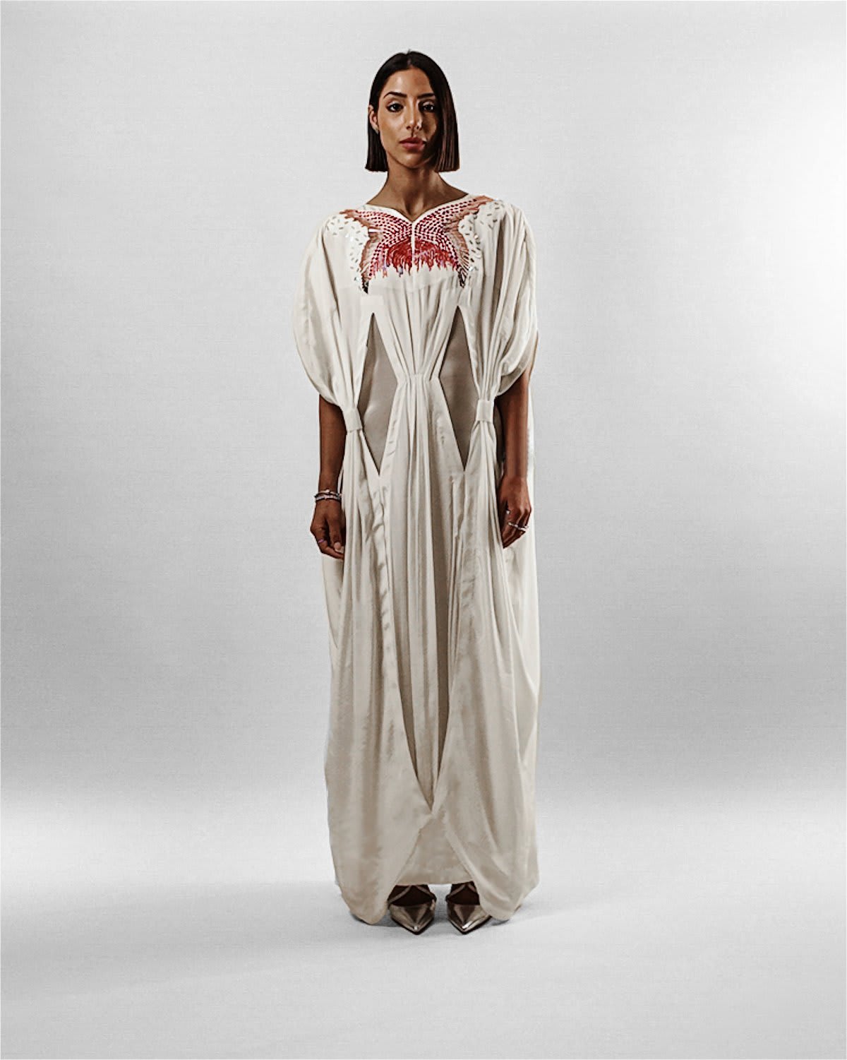 cocora kaftan A full-length studio portrait of a woman with a sleek, dark bob haircut wearing the Cocora Kaftan by Tanisha Rada. The floor-length garment is crafted from a fluid, cream-colored silk with an intricate red and gold butterfly-shaped embroidery across the chest. It features draped, cape-like sleeves and unique geometric fabric overlays that create a structured, multi-dimensional silhouette against a neutral beige background. front